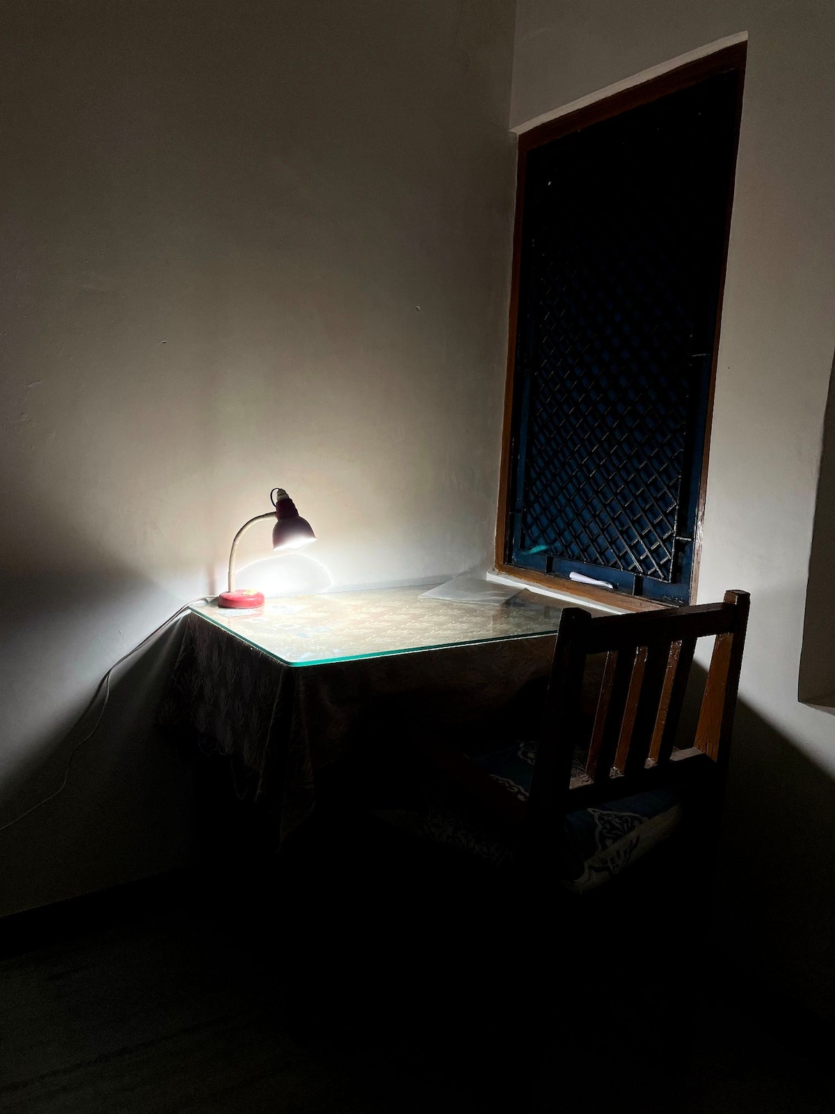 A simple workspace is illuminated by a desk lamp on a table. A wooden chair is positioned at the desk, which is partially visible against a wall. Light filters through a window with a security grille, creating a contrast with the darker room.