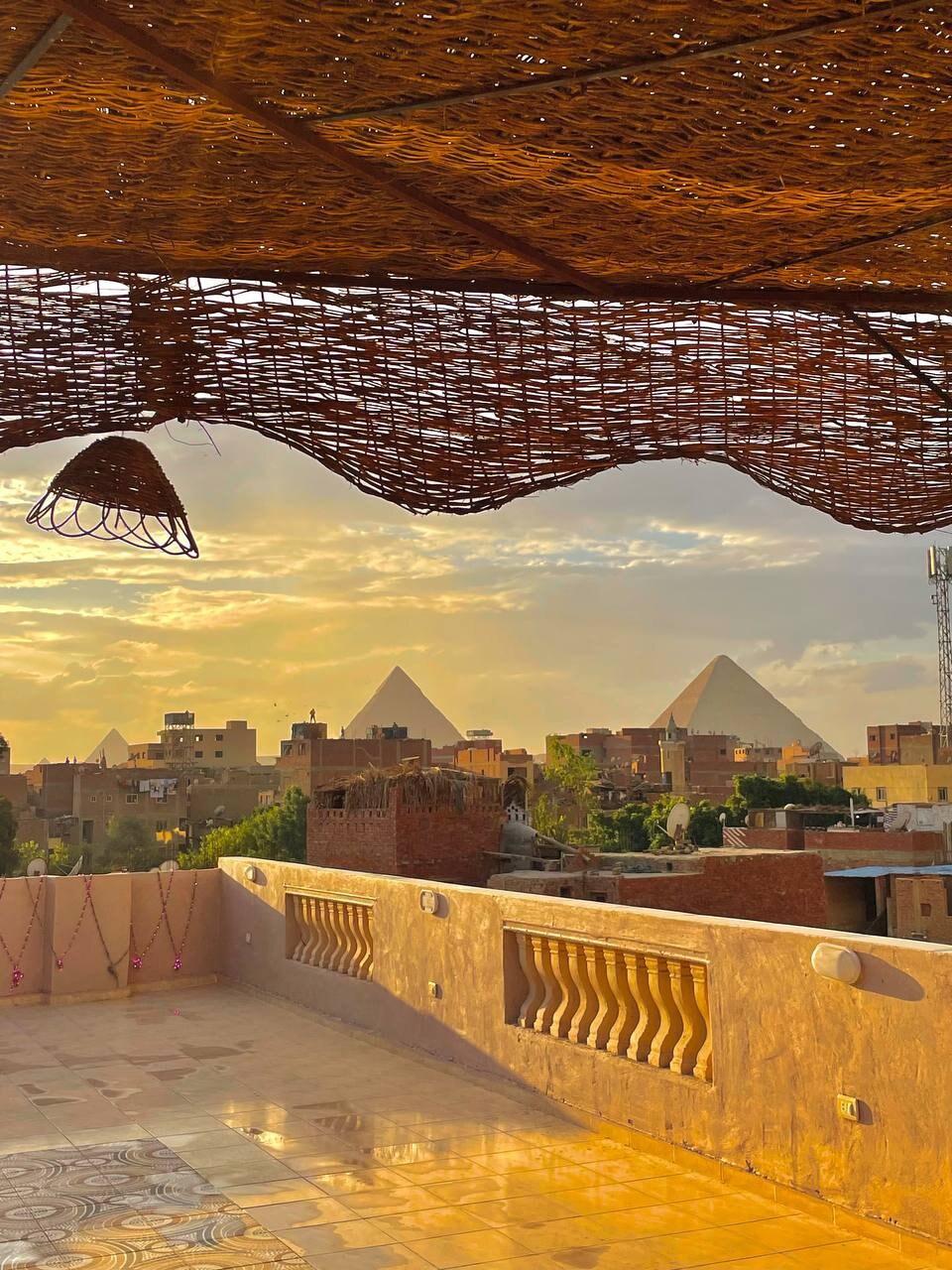 A panoramic view reveals the iconic pyramids against a sunset sky, framed by a woven roof structure. The terrace features a smooth tiled surface, complemented by a decorative railing. Soft lighting is provided by a hanging lantern, enhancing the serene ambiance of the outdoor space.