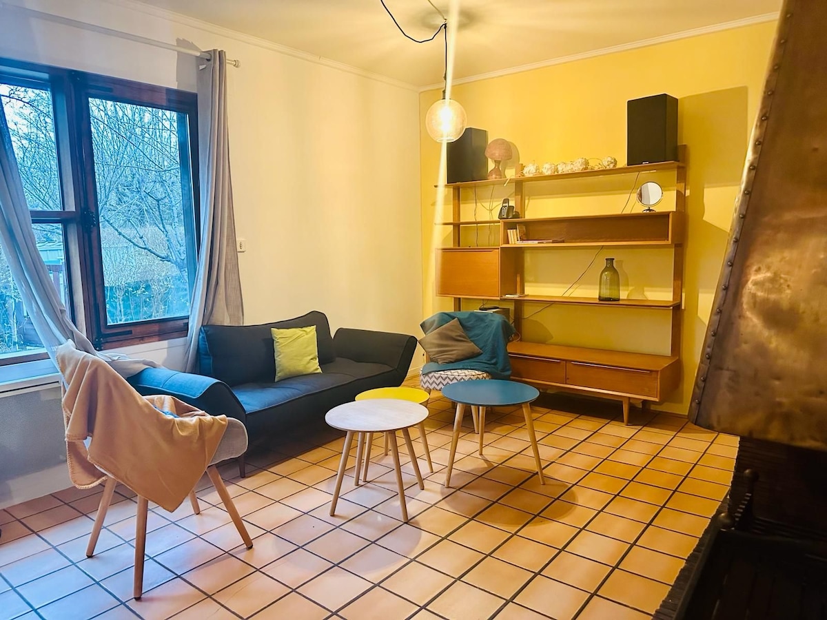 A living area is presented with a modern sofa featuring colorful cushions. Two round coffee tables in varied colors complement a wooden shelving unit, which displays decor items. A large window allows natural light, and light-toned tiles enhance the spacious appearance.