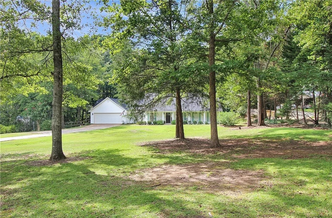 A serene view of the countryside home is framed by tall trees, displaying a well-maintained front lawn. The residence features a white exterior and a spacious driveway leading to an attached garage.