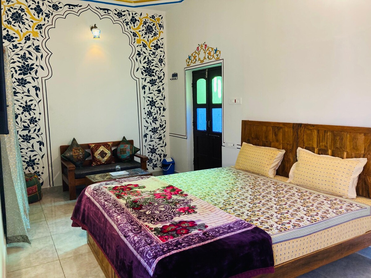 A spacious sleeping area features a large bed adorned with colorful bedding and a decorative throw. Intricate floral designs on the wall are complemented by a cozy seating area with colorful cushions. Natural light enters through a window, creating a serene environment.