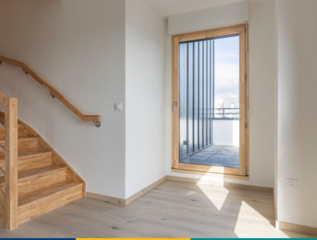 A bright corner of a room showcases a wooden staircase leading to the upper level. Large glass doors provide access to a small balcony, allowing natural light to enter the space. Neutral-colored walls and light wooden flooring create a calming environment.