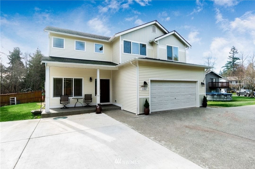 The spacious two-story home showcases a welcoming front porch with seating. The exterior features light-colored siding, large windows, and a double garage. A well-maintained lawn surrounds the property, with additional landscaping visible on the side.