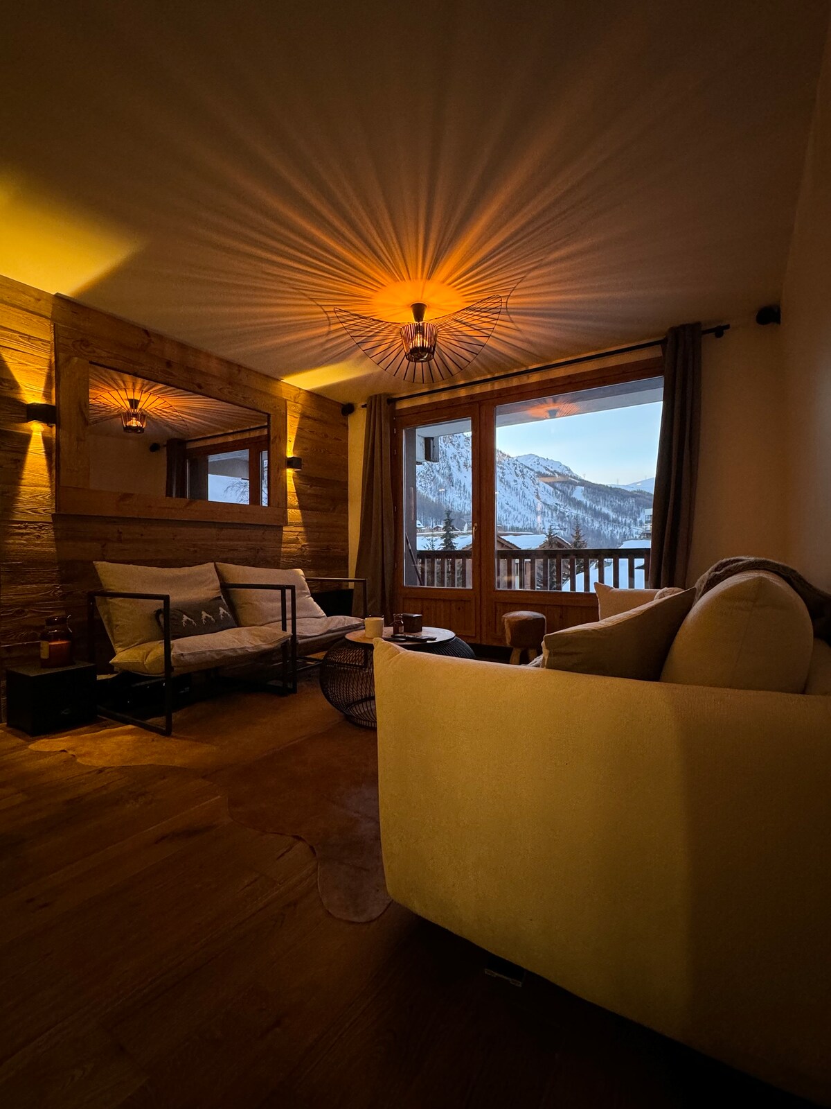 A cozy lounge area is illuminated by warm lighting, featuring a comfortable sofa and armchairs arranged thoughtfully for seating. Large windows provide views of the surrounding mountains, with natural light softening the interior ambiance.