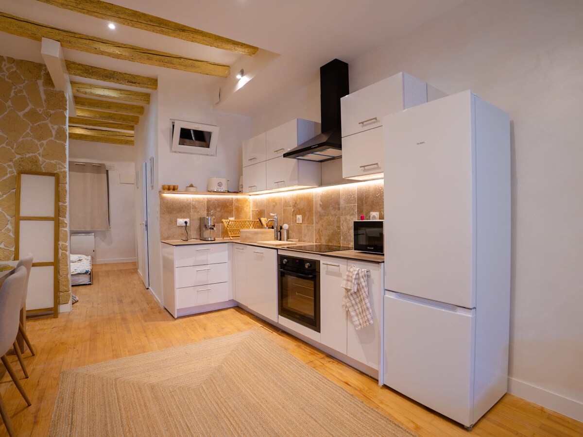 An open kitchen area features modern appliances, including a refrigerator and an oven, arranged along a well-lit countertop. Soft lighting highlights the textured backsplash, and wood beams are visible above, contributing to the space's inviting layout.