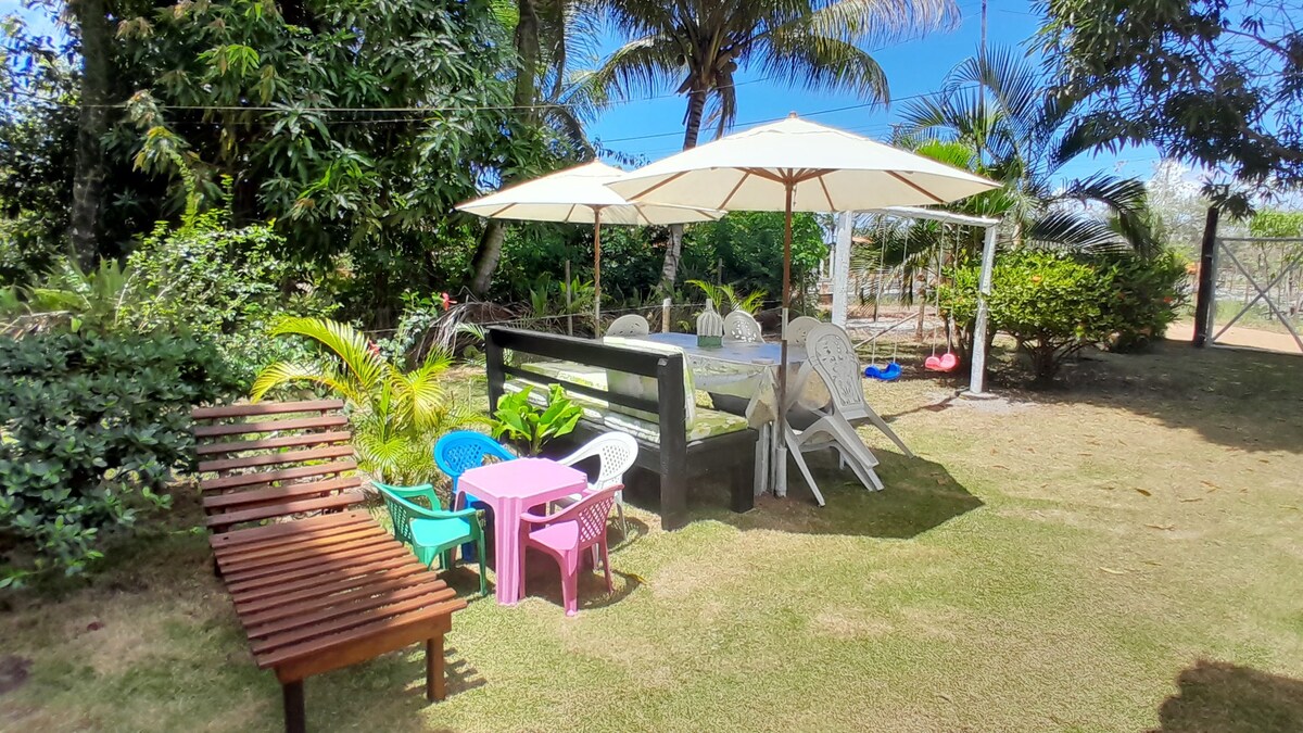 An inviting outdoor area features shaded seating under large umbrellas, surrounded by lush greenery. A wooden bench is present alongside colorful plastic chairs and a small table for children, providing a welcoming space for relaxation and gatherings.