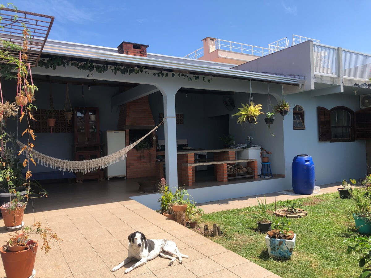 An outdoor space features a shared garden with various plants, a hammock hanging under a shaded area, and an outdoor kitchen equipped with a stove and sink. A friendly dog rests on the patio, while a large blue water barrel is visible nearby.