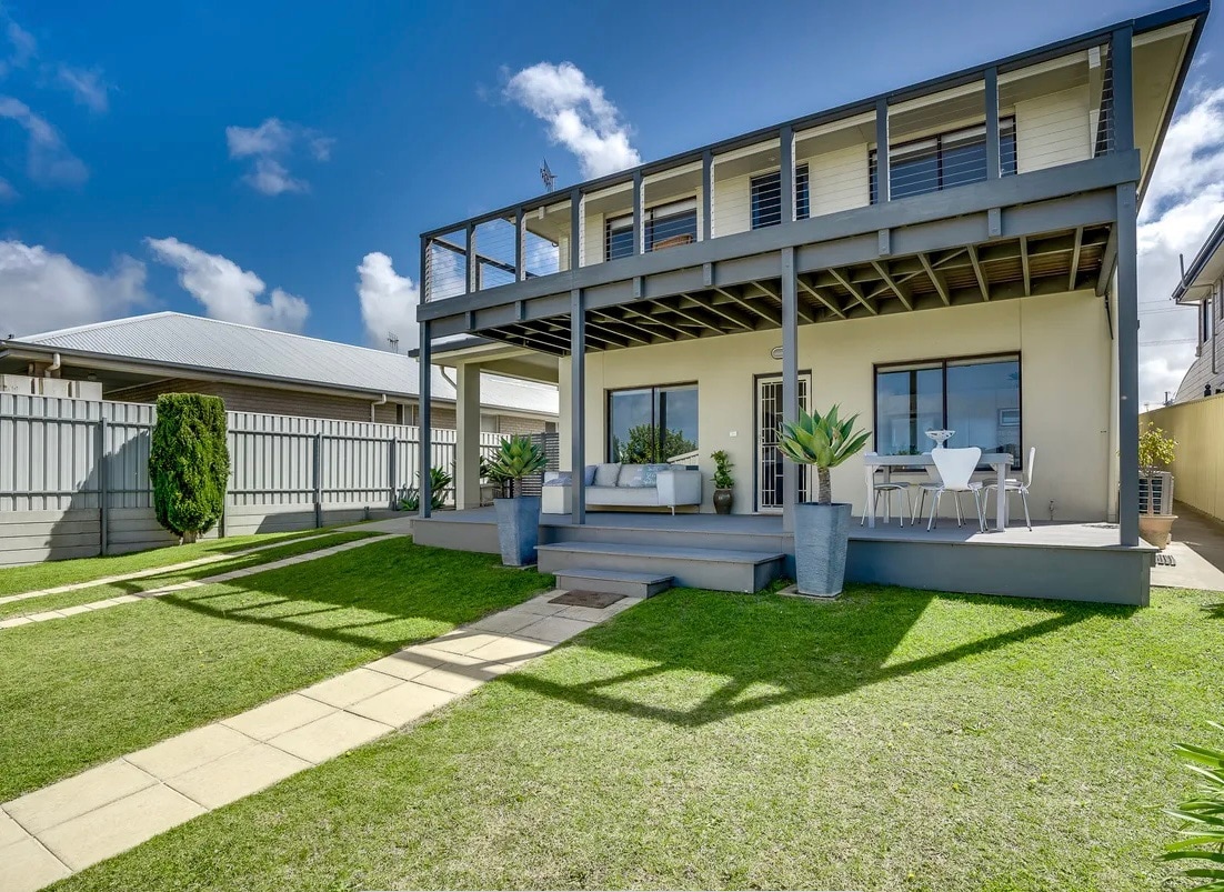 A modern two-story holiday home is featured with a spacious deck area furnished with seating and a dining table. The well-manicured lawn and stone pathway lead to the entrance, while large windows allow natural light to fill the interior.