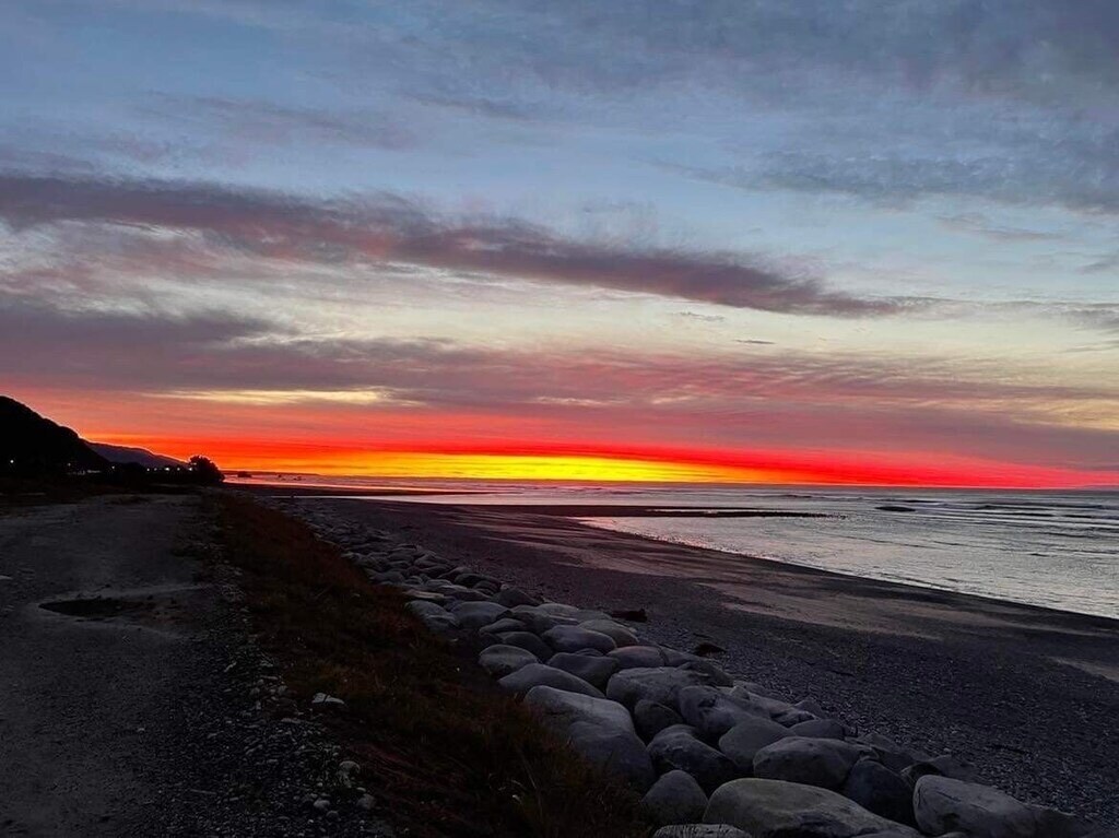 A serene coastal scene captures a vivid sunset over the horizon, with hues of orange, pink, and purple melting into the sky. Pebbles line the beach, and the calm water reflects the colorful display, providing a peaceful backdrop for relaxation.