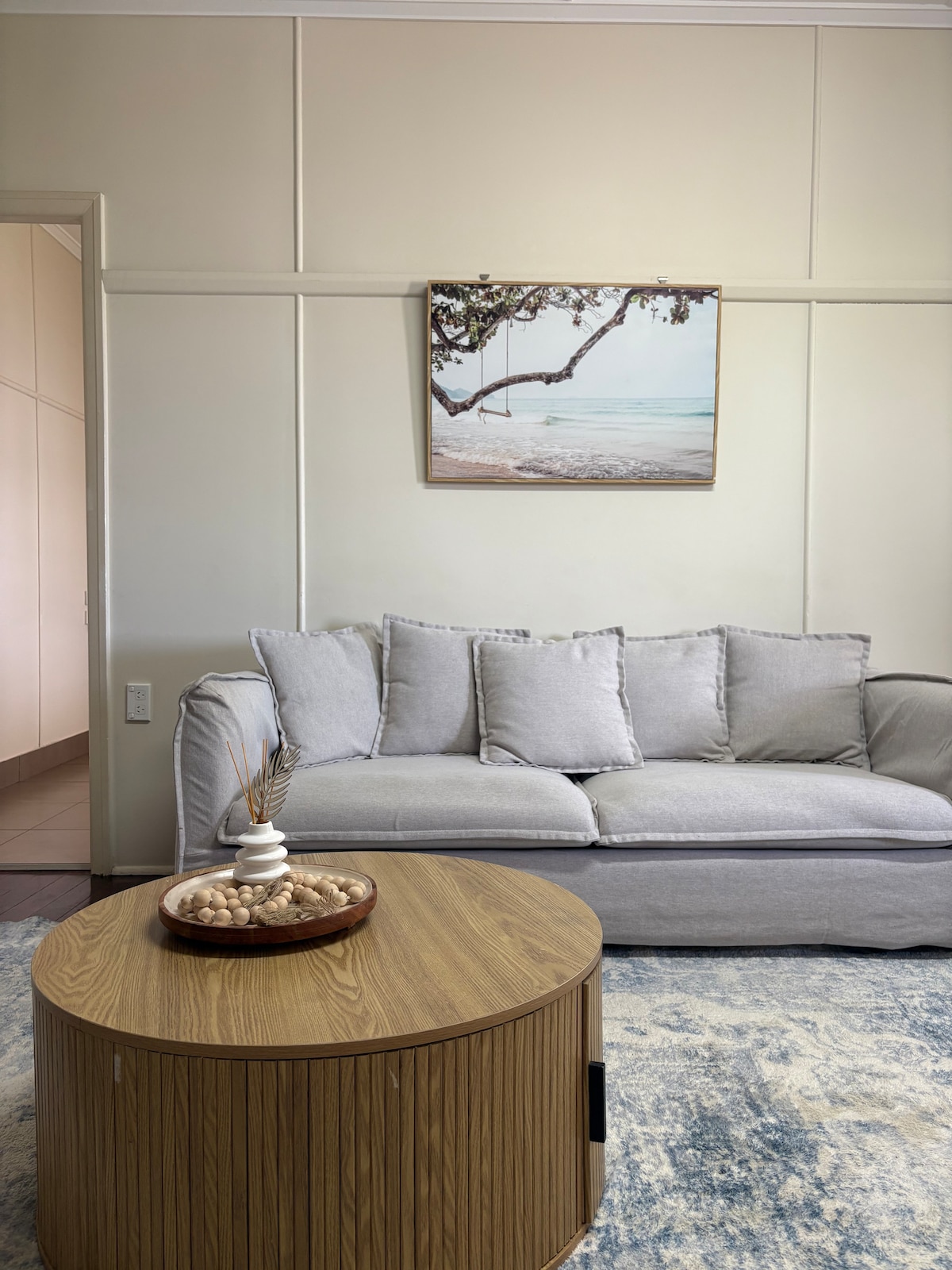 A light-colored wall frames a cozy seating area featuring a soft gray sofa. Above the sofa, a beach-themed artwork adds a coastal touch. A round wooden coffee table is placed at the center, showcasing a decorative bowl with natural elements.