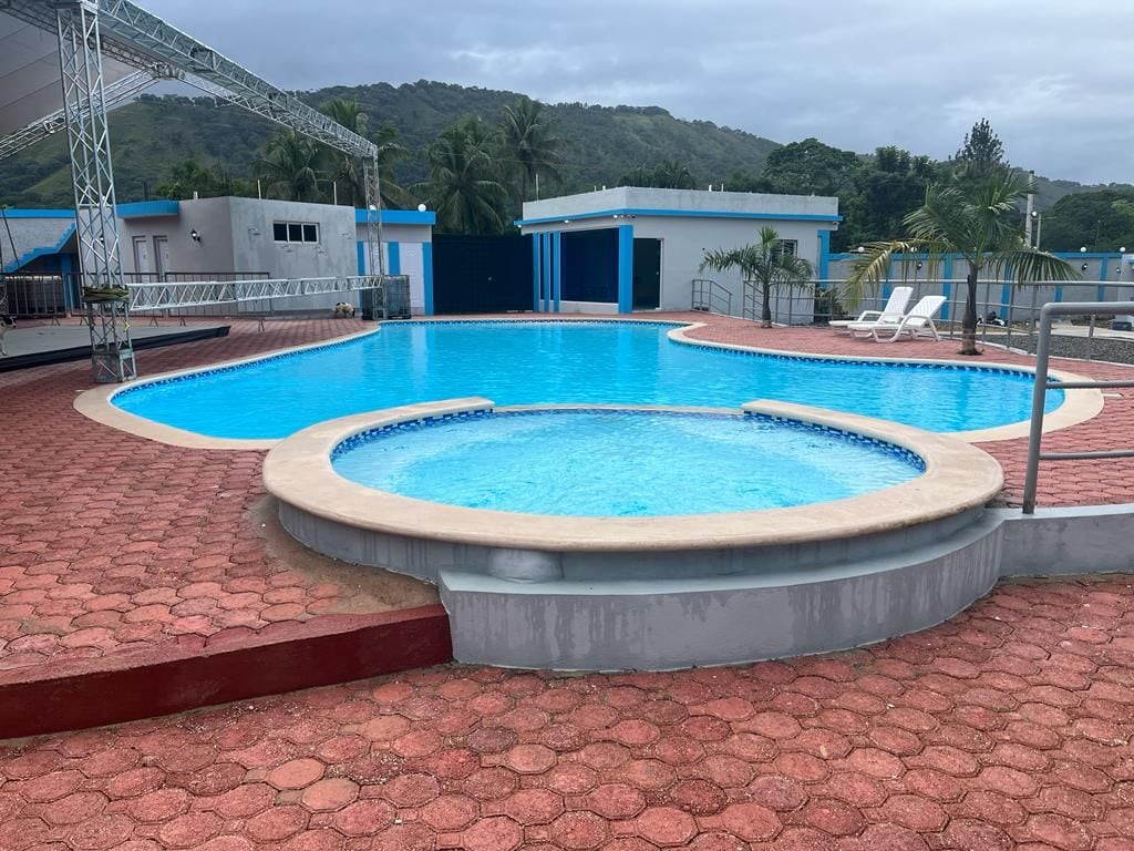 A large swimming pool with clear blue water is surrounded by a red-brick deck. A smaller circular pool is adjacent, promoting relaxation. Several white lounge chairs are positioned around the area, with a backdrop of lush green hills and palm trees.