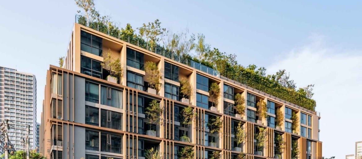 A modern apartment building features multiple levels with large glass windows, allowing natural light to fill the interior. Lush greenery is placed on the roof and balconies, enhancing the building's aesthetic. The surrounding area includes other urban structures, contributing to a vibrant city atmosphere.