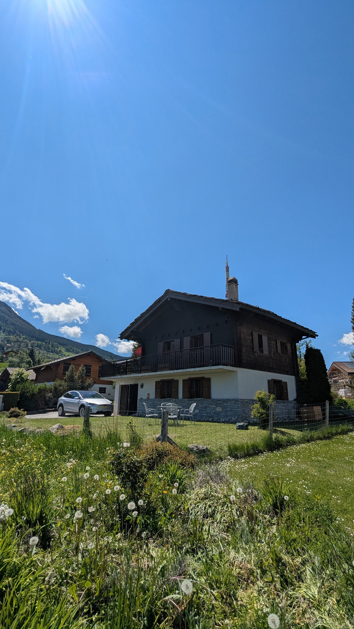 The chalet stands prominently against a clear blue sky, featuring a combination of wood and stone elements. Lush greenery and wildflowers surround the property, with a spacious lawn leading to the entrance. A driveway accommodates a car, and mountains are visible in the background.