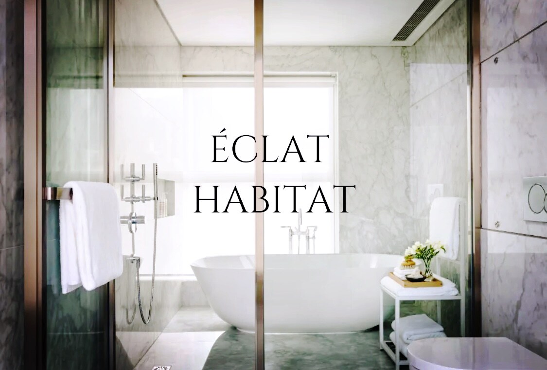 A modern bathroom features a freestanding soaking tub, framed by glass panels. Neutral marble walls and a sleek stone floor contribute to a clean aesthetic. Soft towels are neatly arranged on a chrome rack, while a small plant adds a touch of greenery on a minimalist side table.