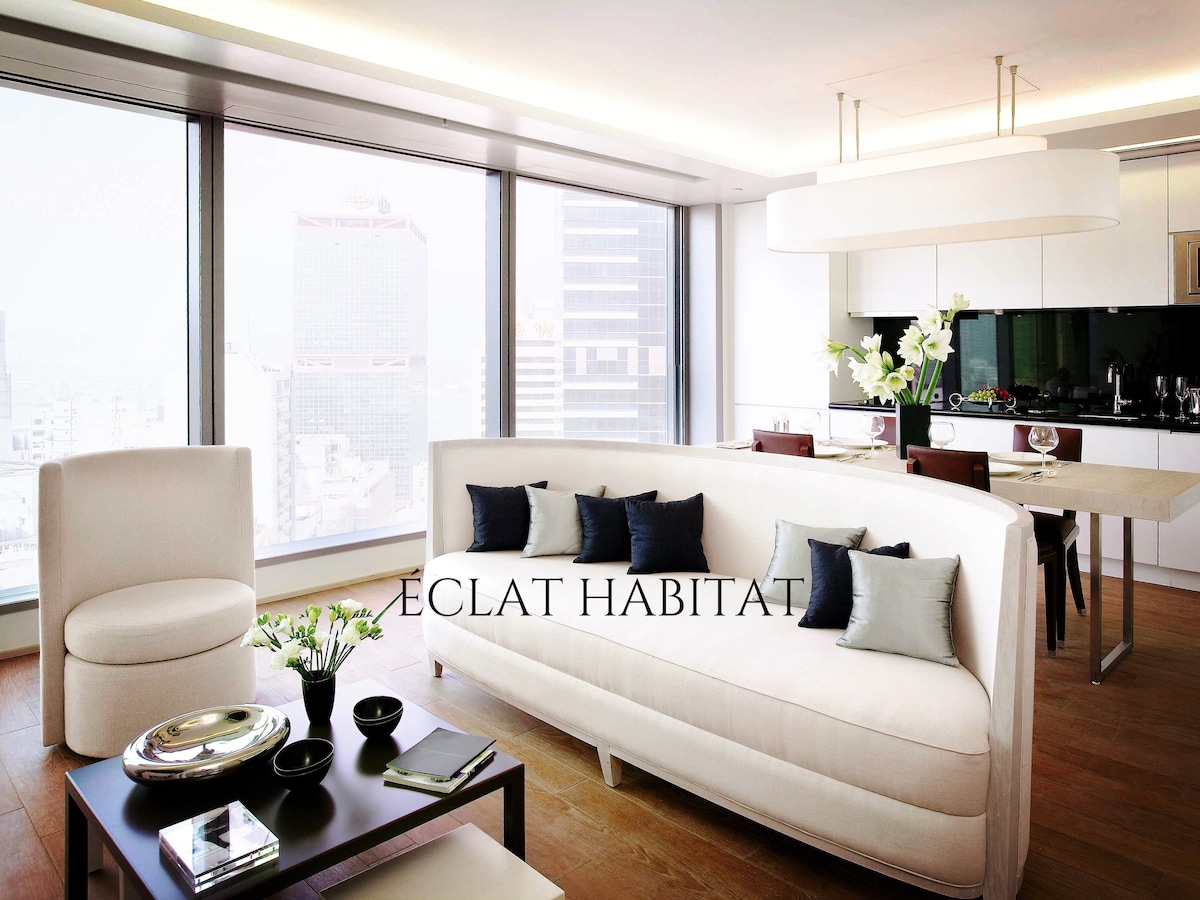 A stylish living area is presented with a large, curved white sofa adorned with dark blue and light green cushions. A dining table is visible in the background, accompanied by modern chairs. Floor-to-ceiling windows offer views of the surrounding cityscape.