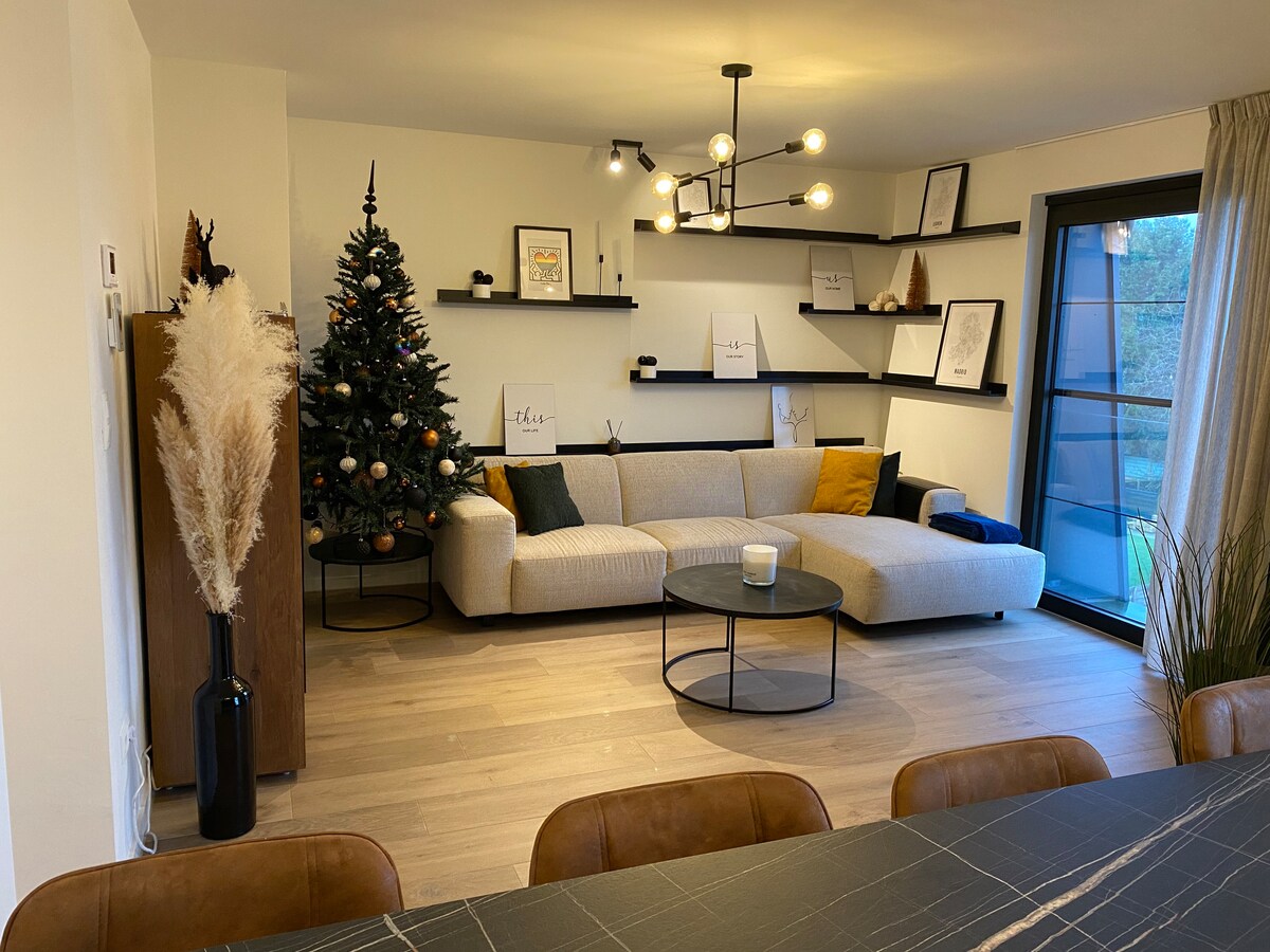 A modern living area features a large, light-colored sectional sofa and a round coffee table on warm-toned flooring. A Christmas tree adorned with decorations stands in the corner. Minimalist shelves showcase framed art. A large window offers views of the outside, contributing to the decor.