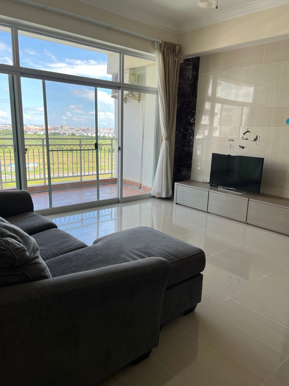A modern living area features a comfortable gray sofa positioned near floor-to-ceiling glass doors, allowing for natural light and views of the surrounding greenery. A flat-screen TV is placed on a sleek entertainment unit against a stylish wall.