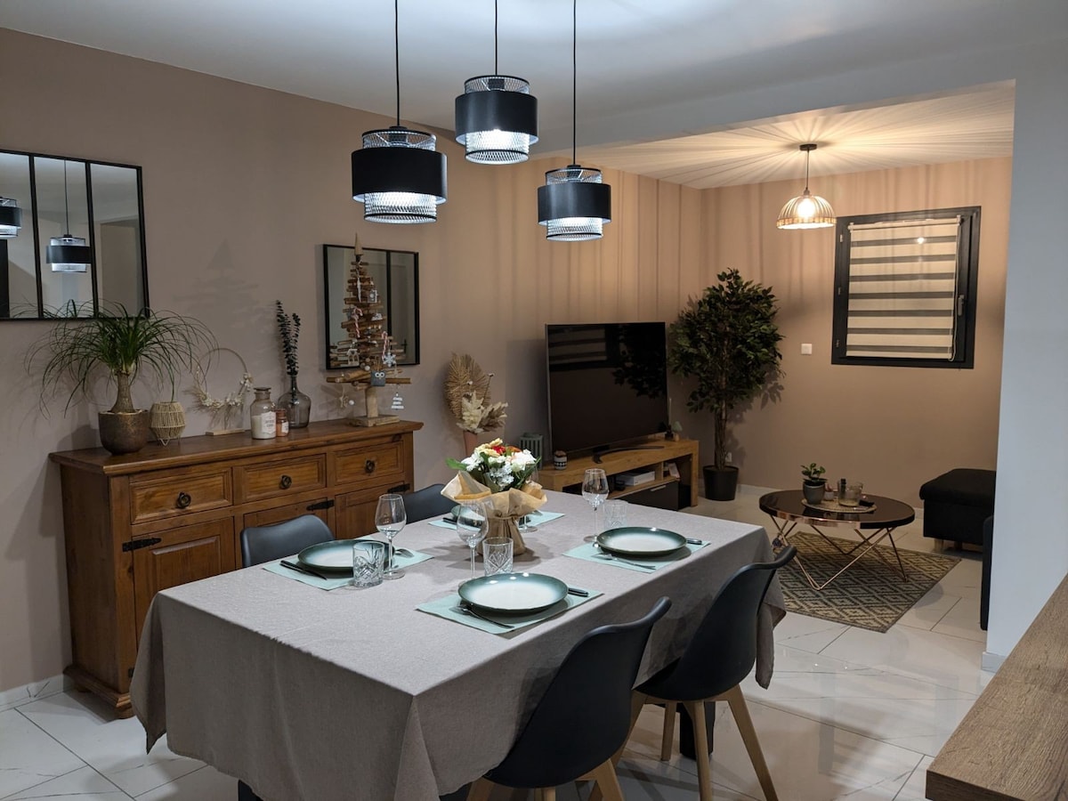 A dining area features a light brown wall and a spacious table set for four, with elegant tableware and a floral centerpiece. Modern lighting fixtures hang from the ceiling, illuminating the space. A wooden sideboard stands nearby, complemented by decorative plants and artwork.