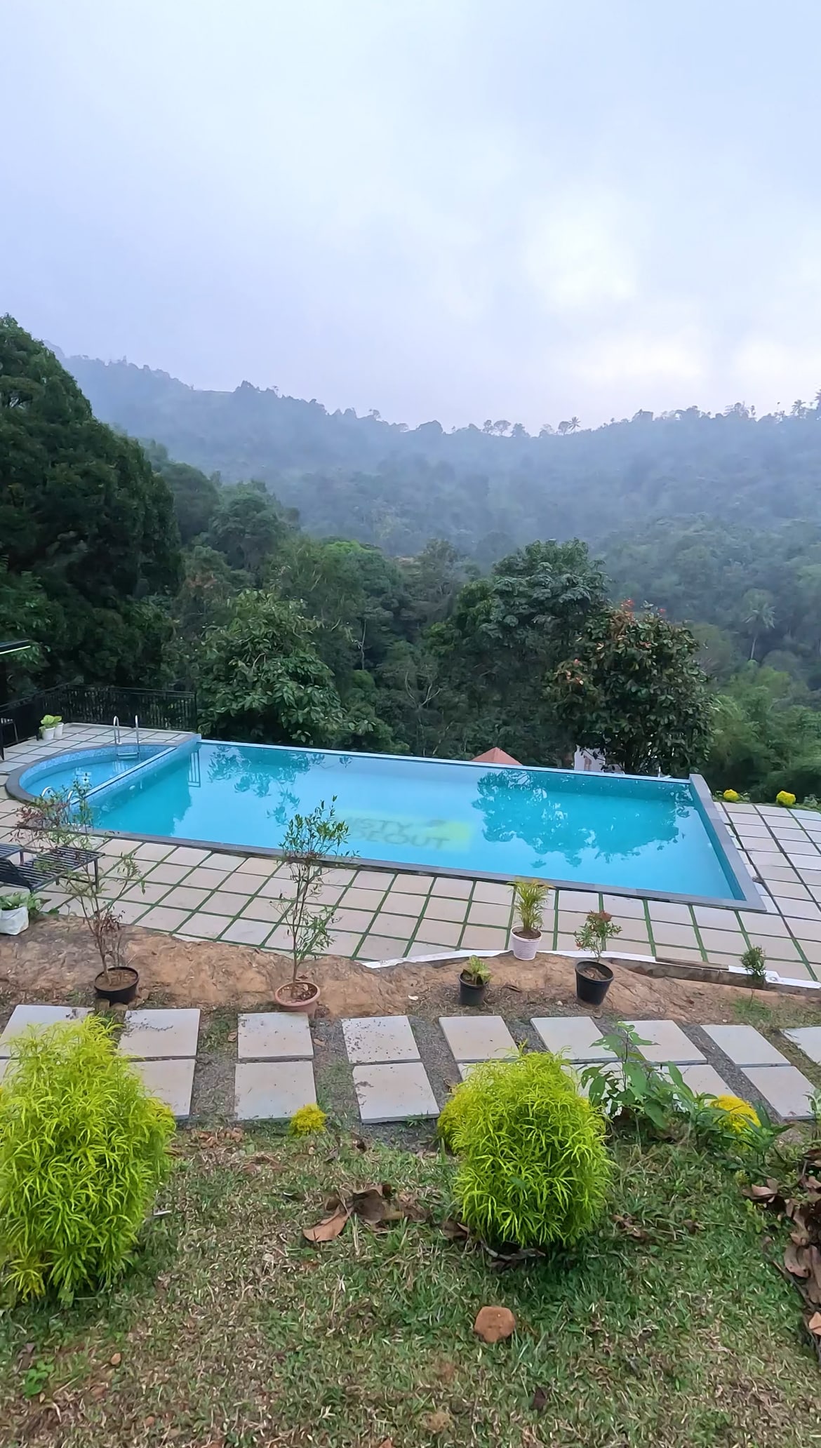 A serene outdoor pool is framed by lush greenery and smooth stone pathways. The surrounding hills are shrouded in mist, creating a tranquil natural setting. Potted plants are arranged neatly along the edge, enhancing the peaceful ambiance.