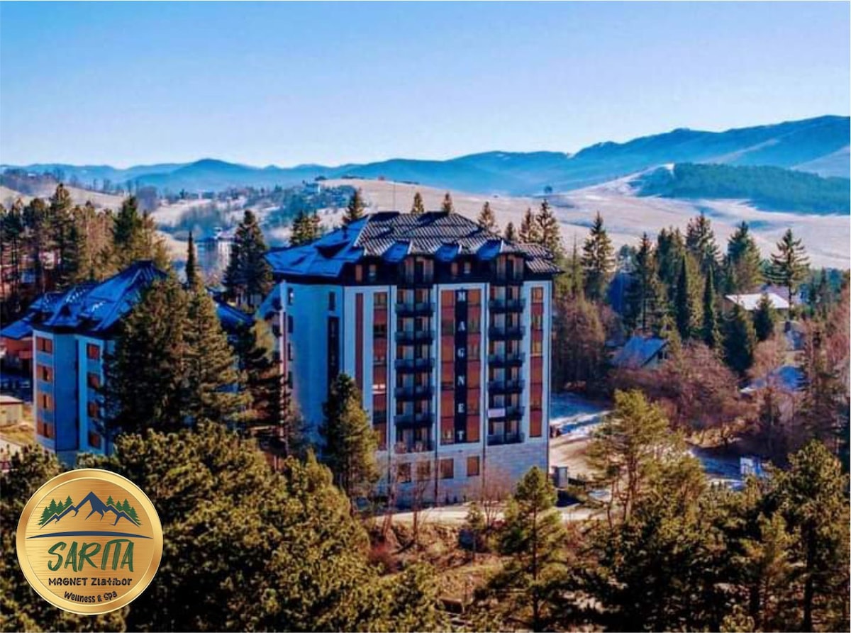 The apartment building is nestled among tall trees, with a backdrop of rolling hills and distant mountains. The structure features multiple balconies, large windows, and a combination of stone and wood elements, blending with the natural surroundings.
