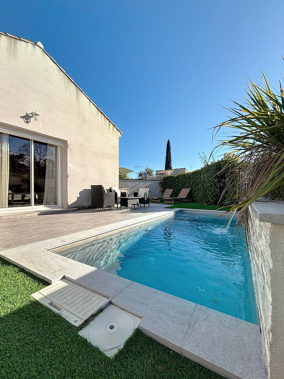 A serene outdoor space is depicted, featuring a sparkling pool beside a wooden deck. Several lounge chairs are positioned for sunbathing. Lush greenery frames the area, enhancing the peaceful setting. Large windows of the villa reflect the clear blue sky, inviting natural light indoors.