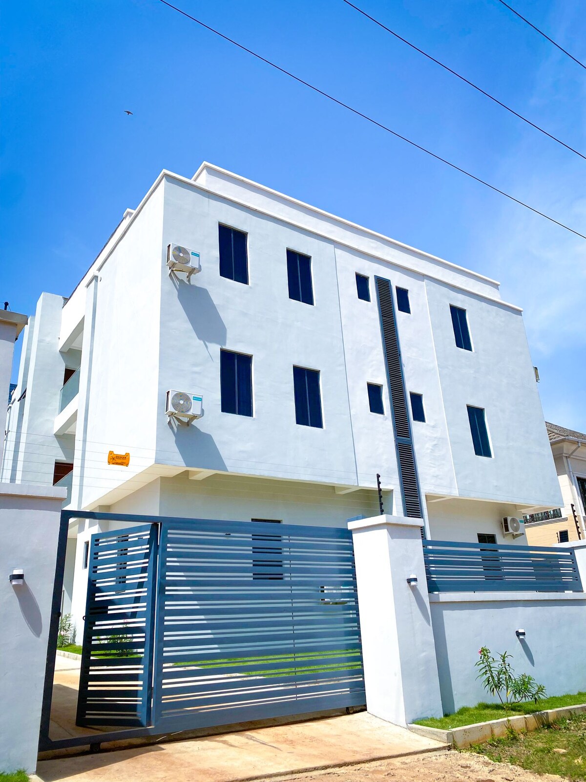 A modern three-story building is presented, featuring a pristine white exterior complemented by several evenly spaced windows. A secure gate leads to the entrance, and the surrounding area includes well-maintained greenery, contributing to a welcoming first impression.
