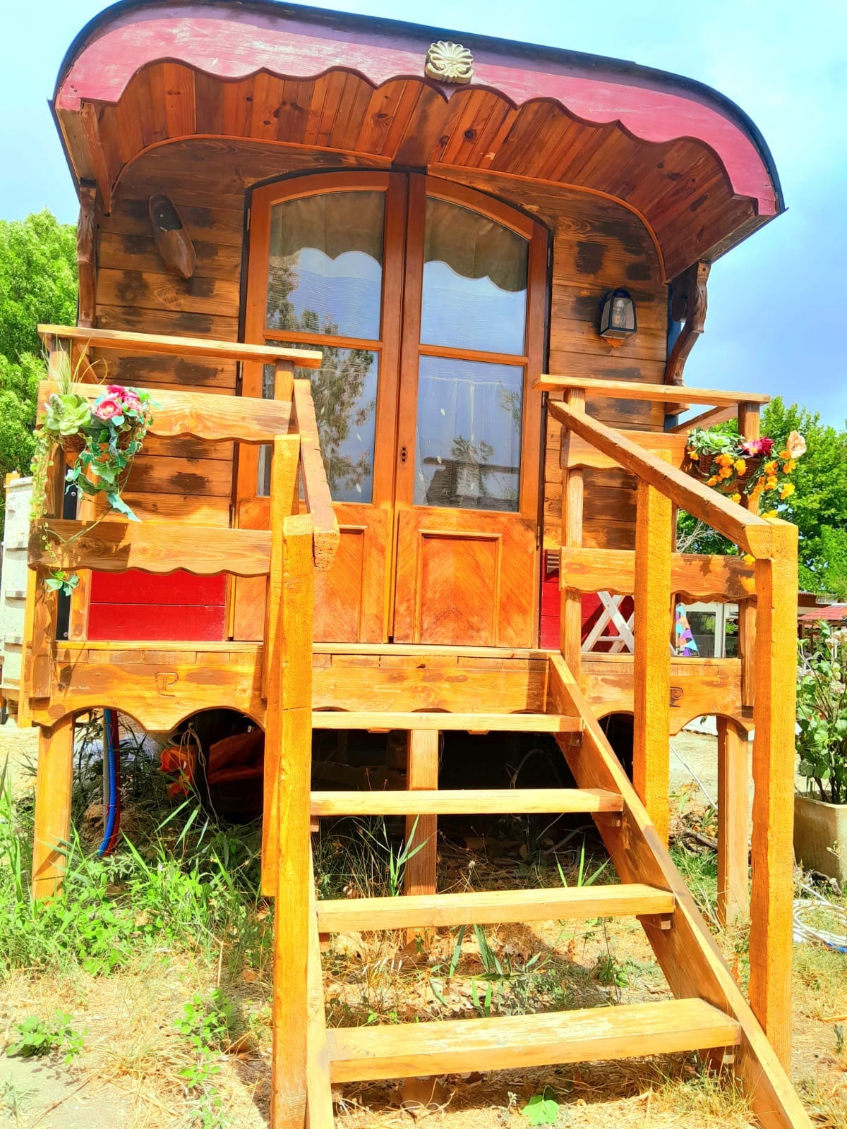 The exterior of a charming wooden roulotte is shown, featuring a covered porch with steps leading up. Decorative flowers adorn the entrance, and large windows allow natural light to filter in. The warm wood tones are complemented by a red awning above the door.