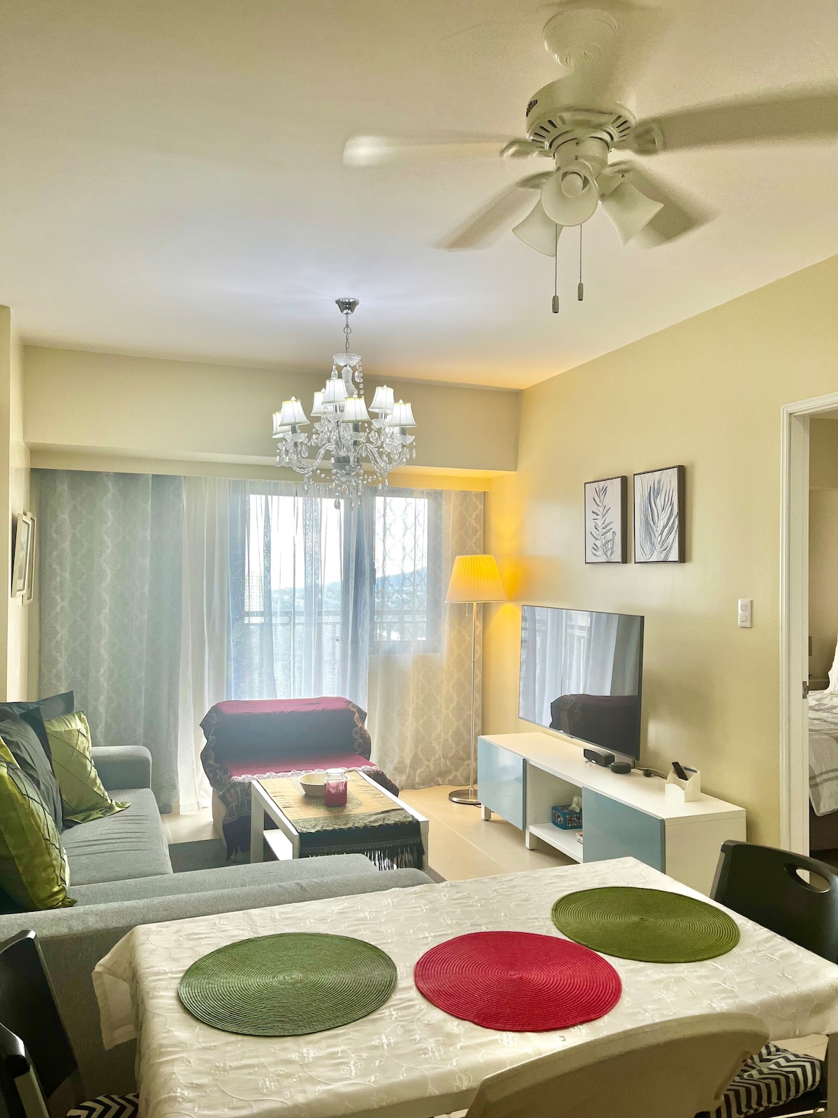 A well-lit sitting area features a ceiling fan and a stylish chandelier. A comfortable sofa is adorned with colorful cushions, while a TV stand holds a 50-inch SmartTV. A dining table is set with red and green placemats. Sheer curtains allow natural light to filter in.