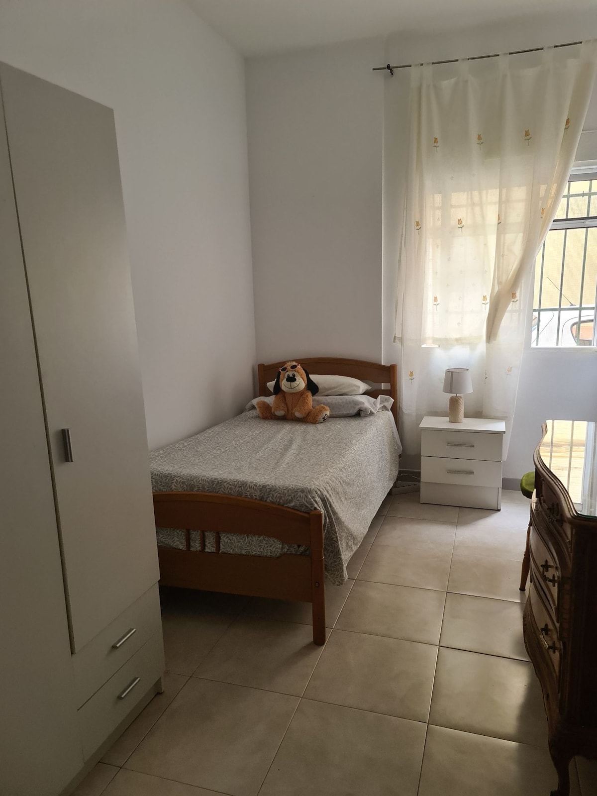 A compact bedroom features a single bed with a light gray blanket and a plush dog toy placed on the pillows. A small nightstand with a lamp is positioned next to the bed, and a wardrobe is visible on one side. Natural light filters through sheer curtains.