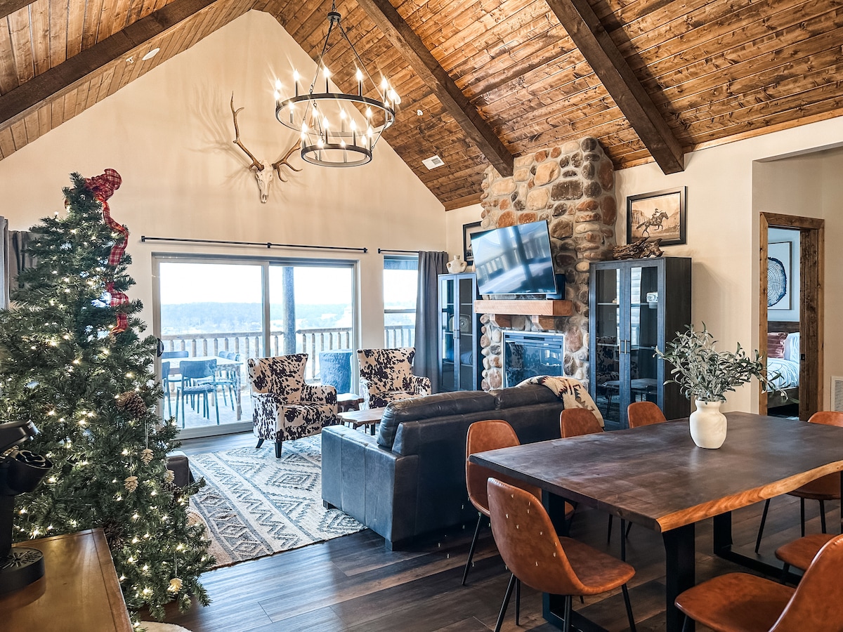 A spacious living area features high, vaulted ceilings and wooden beams. A stone fireplace is positioned nearby, and large glass doors provide access to a deck with a lake view. Comfortable seating includes patterned chairs and a dark leather sofa, surrounded by a beautifully decorated Christmas tree.