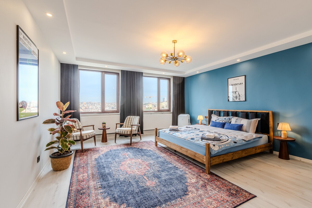 A spacious bedroom features a wooden bed dressed in blue linens, accompanied by a patterned rug. Two accent chairs sit beside a potted plant, while a nightstand holds a lamp. Large windows offer a view of the city, with soft lighting highlighting the room's decor.