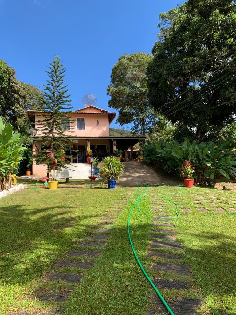 A charming house is framed by lush greenery and colorful planters. A pathway lined with stones leads toward the entrance, with a vibrant blue sky visible above. The two-story building is partially shaded by large trees, creating a serene outdoor environment.