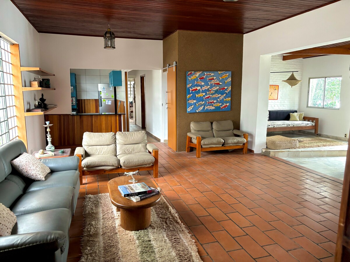 An open living area features a combination of comfortable seating options, including two sofas and a coffee table. A colorful artwork is displayed on the wall, while natural light filters through the large windows, highlighting the warm, earthy tones of the floor and furniture.