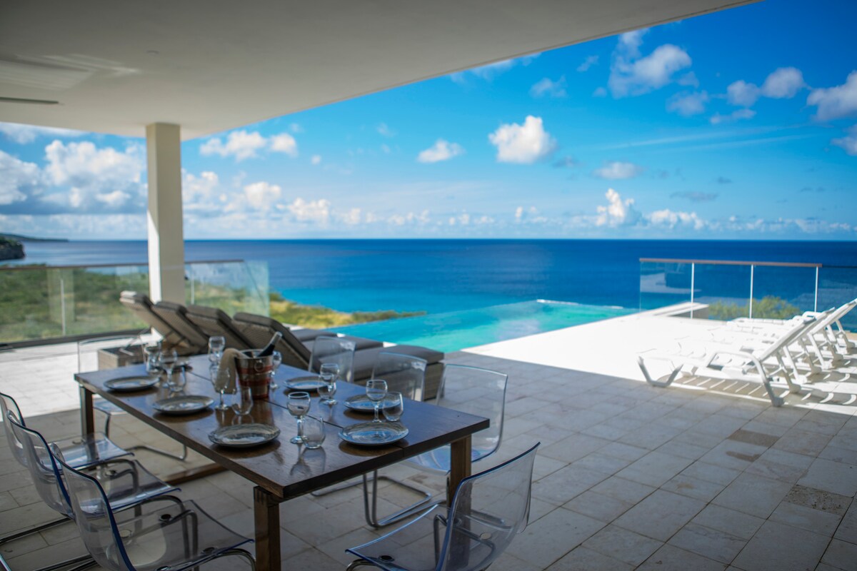An outdoor dining area features a wooden table set with plates and glasses, facing panoramic views of the ocean. Clear skies and turquoise waters can be seen beyond the glass railing. Lounge chairs are positioned nearby on the terrace, inviting relaxation.