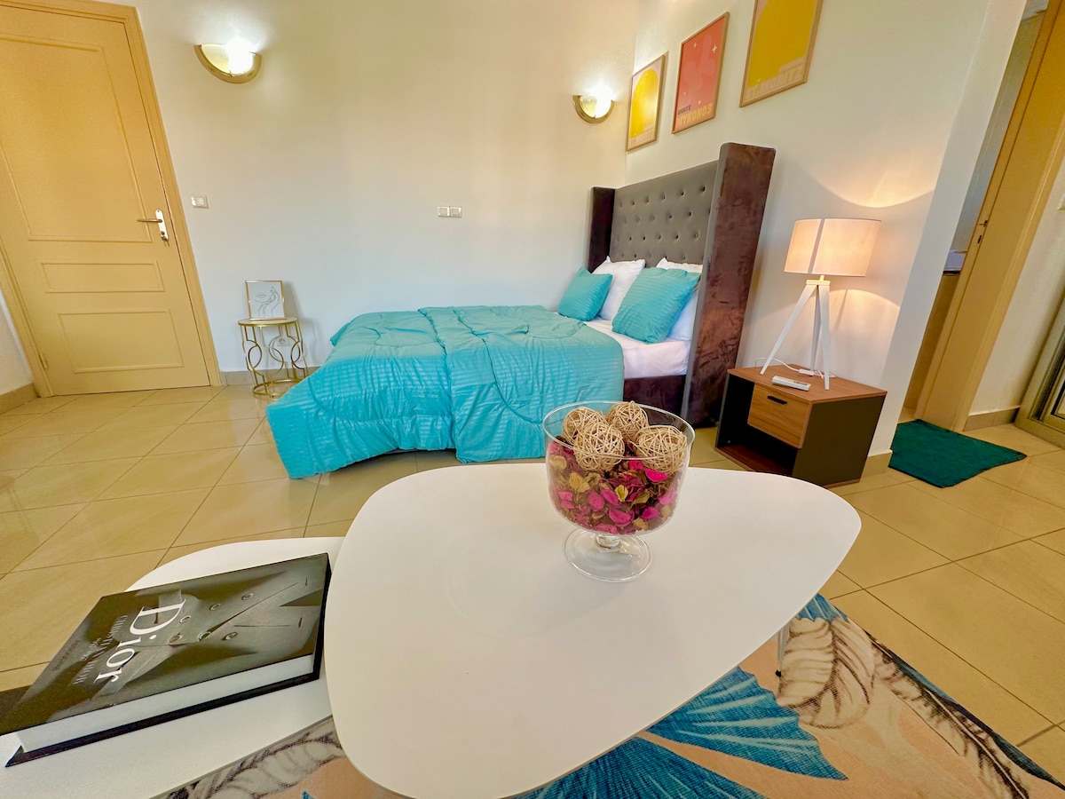 A cozy bedroom is presented with a padded headboard behind a spacious bed covered in a turquoise quilt. A small nightstand and a lamp are positioned beside the bed. A round coffee table sits in front of a decorative arrangement of natural materials.