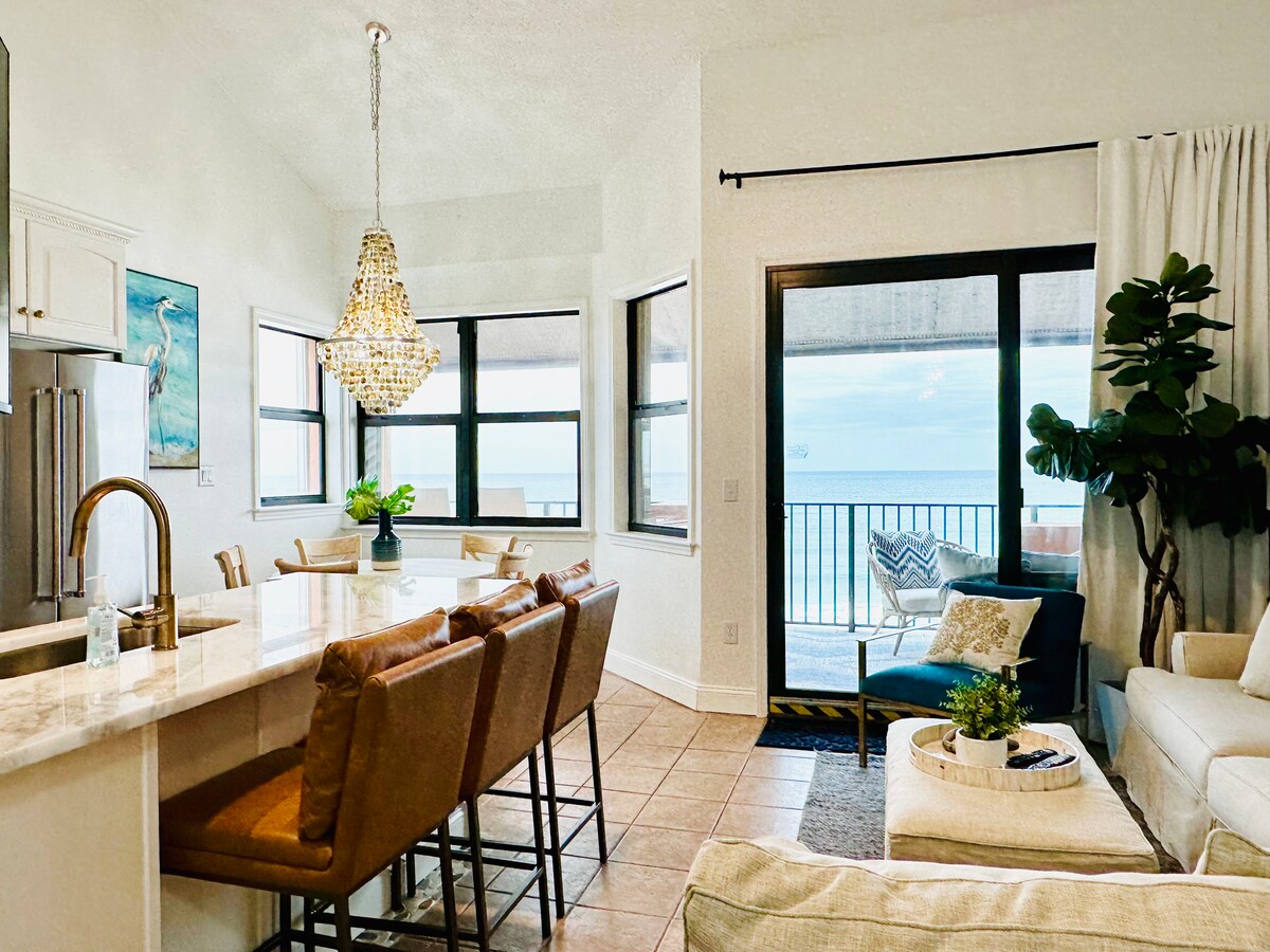 An open living space showcases a large dining table with upholstered chairs, positioned near expansive windows offering views of the beach. A comfortable seating area is visible, complementing a light-colored sofa and decorative plants. A modern chandelier hangs above the dining table, enhancing the contemporary feel.