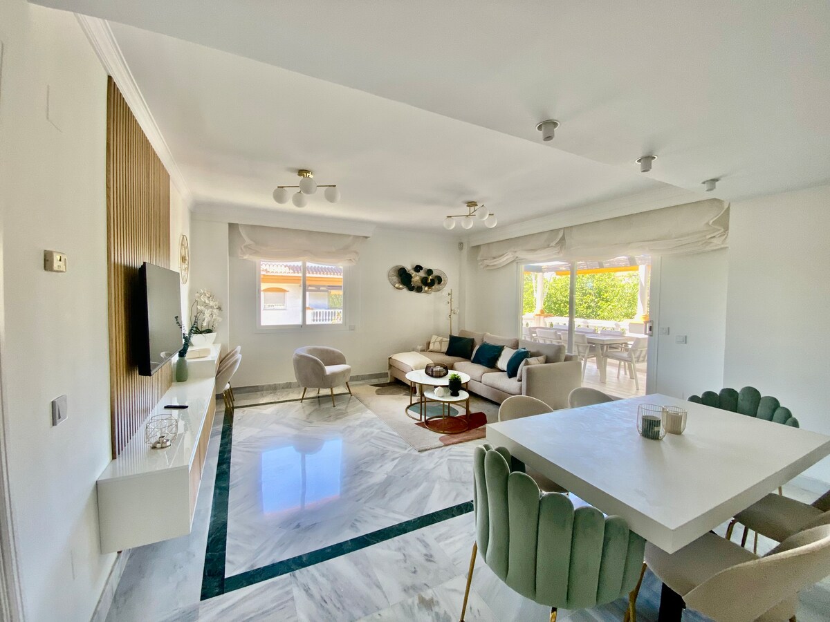 A bright living space features a light-toned sofa arranged around a central area rug. A dining table with plush chairs is positioned nearby. Large windows allow natural light to fill the room, revealing views of the outdoor terrace and greenery.