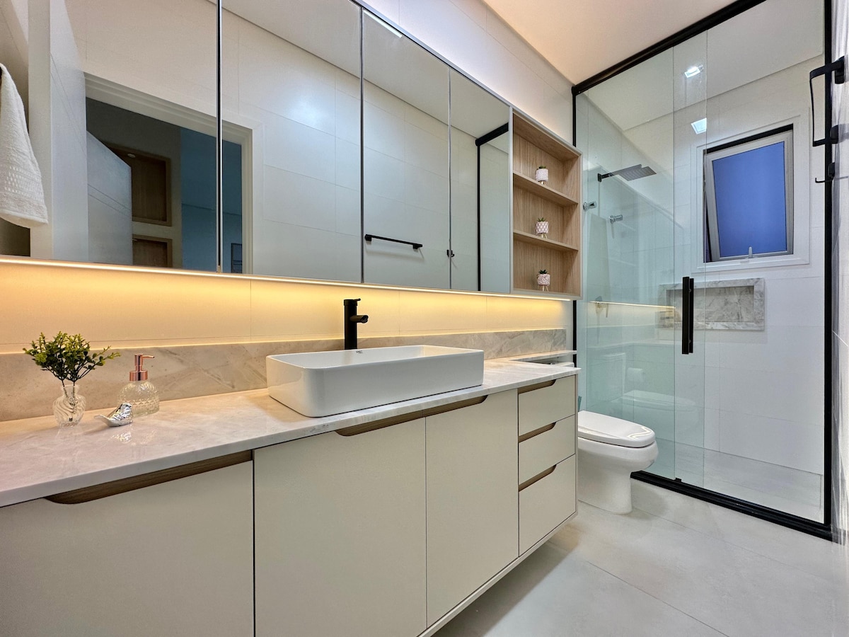 A modern bathroom features a spacious countertop with a rectangular sink, illuminated by warm under-cabinet lighting. A large mirror extends across the wall above, while a glass-enclosed shower occupies one corner. Simple shelves display toiletries, contributing to the organized and sleek design.