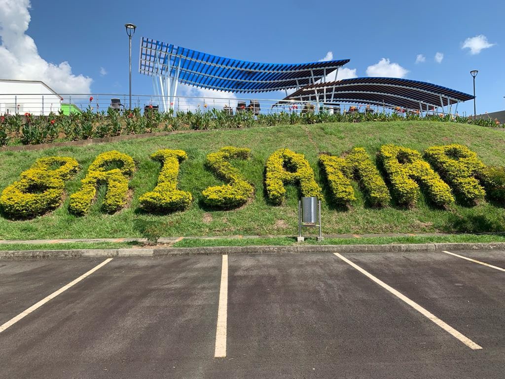 A vibrant landscape is showcased with the name 'BRISAMAR' artistically formed from neatly trimmed hedges. Surrounding greenery enhances the scene, with clear blue skies above and modern architectural elements visible in the background.