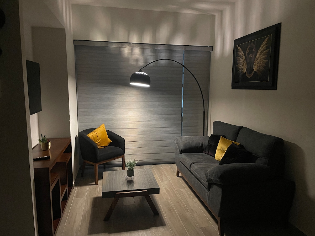 A cozy living room features a dark gray sofa paired with a black armchair accented by a yellow pillow. A floor lamp provides soft lighting above a low wooden table, while a plant adds a touch of greenery. Blinds cover the window, allowing for adjustable light.