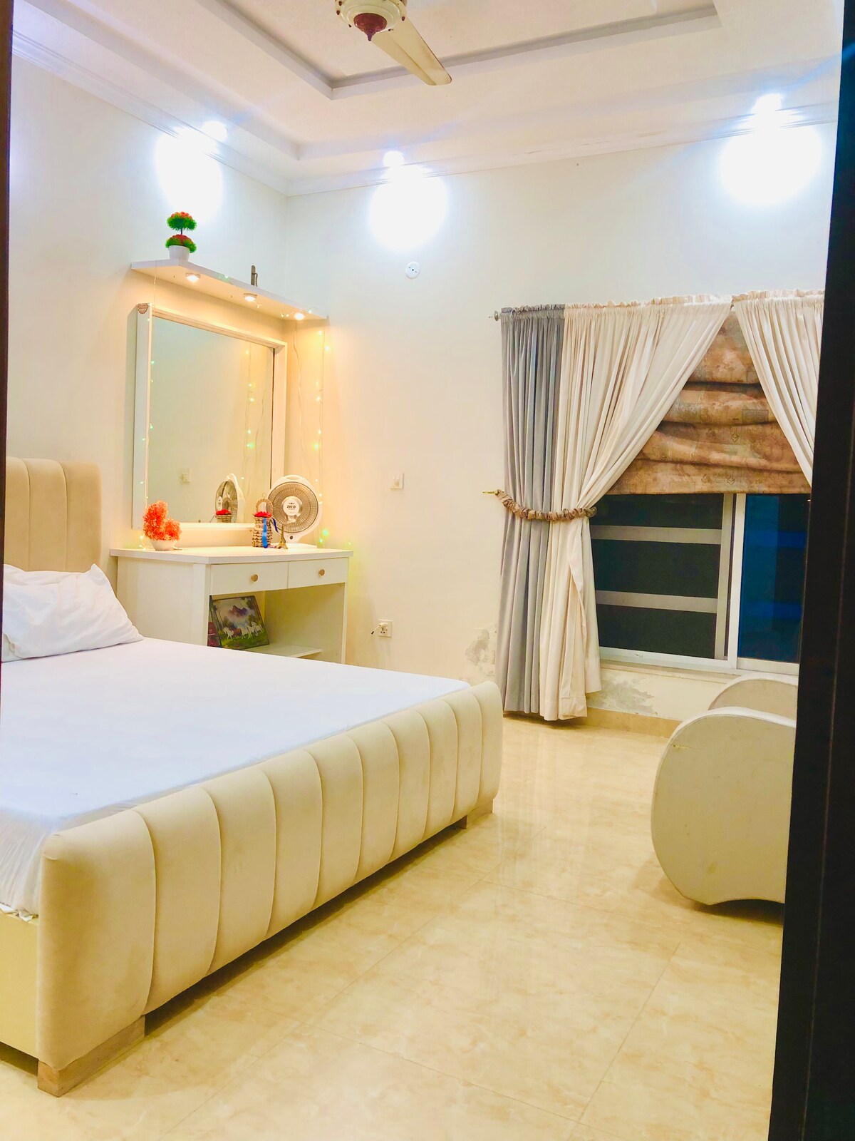 A well-furnished bedroom features a light-colored bed and a modern vanity with decorative lights. Sheer curtains frame a large window, allowing natural light to fill the space. A stylish seating area is present, adding to the room's overall functionality and comfort.