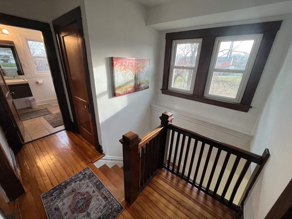 A well-lit hallway features a wooden staircase leading down, accentuated by a small area rug. Large windows allow natural light to fill the space. A vibrant artwork depicting autumn trees hangs on the wall, complementing the rich wooden trims and warm hardwood flooring.