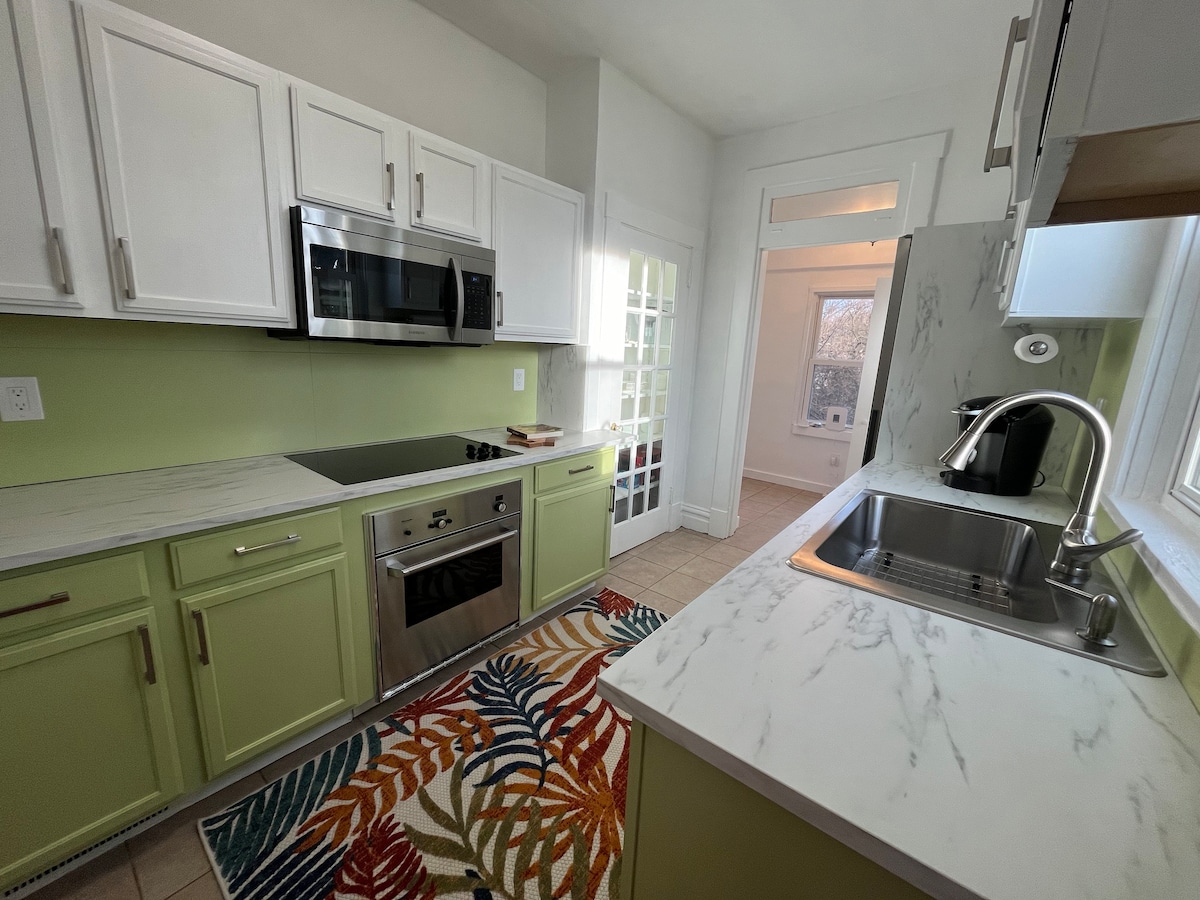 A modern kitchen features white cabinetry with green accents and marble countertops. Stainless steel appliances, including a microwave and oven, are visible. A colorful area rug adds vibrancy, while a window allows natural light to brighten the space.