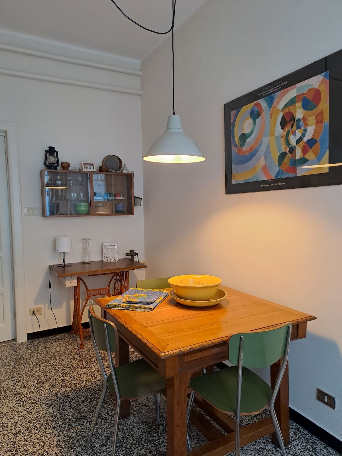 A wooden dining table is set for four, accompanied by green chairs. A bowl sits at the center, and magazines are casually arranged beside it. A wall-mounted shelf displays glassware and decorative items, while a colorful piece of artwork enhances the atmosphere.