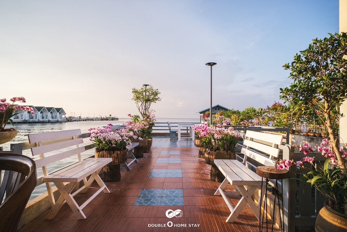 An outdoor terrace features white benches arranged among colorful potted flowers. The space overlooks the water, with soft hues of blue and pink in the sky. Pathways of smooth tiles are bordered by planters, creating a peaceful setting for relaxation.
