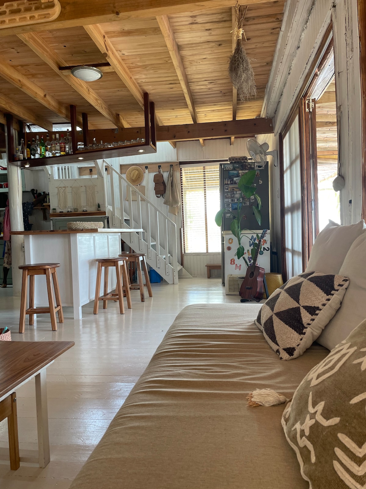 A cozy living space is highlighted by a comfortable sofa adorned with decorative pillows. Light wood beams create an open atmosphere, with a glimpse of the kitchen and dining area in the background. A set of wooden stools is positioned near a minimalist kitchen island.