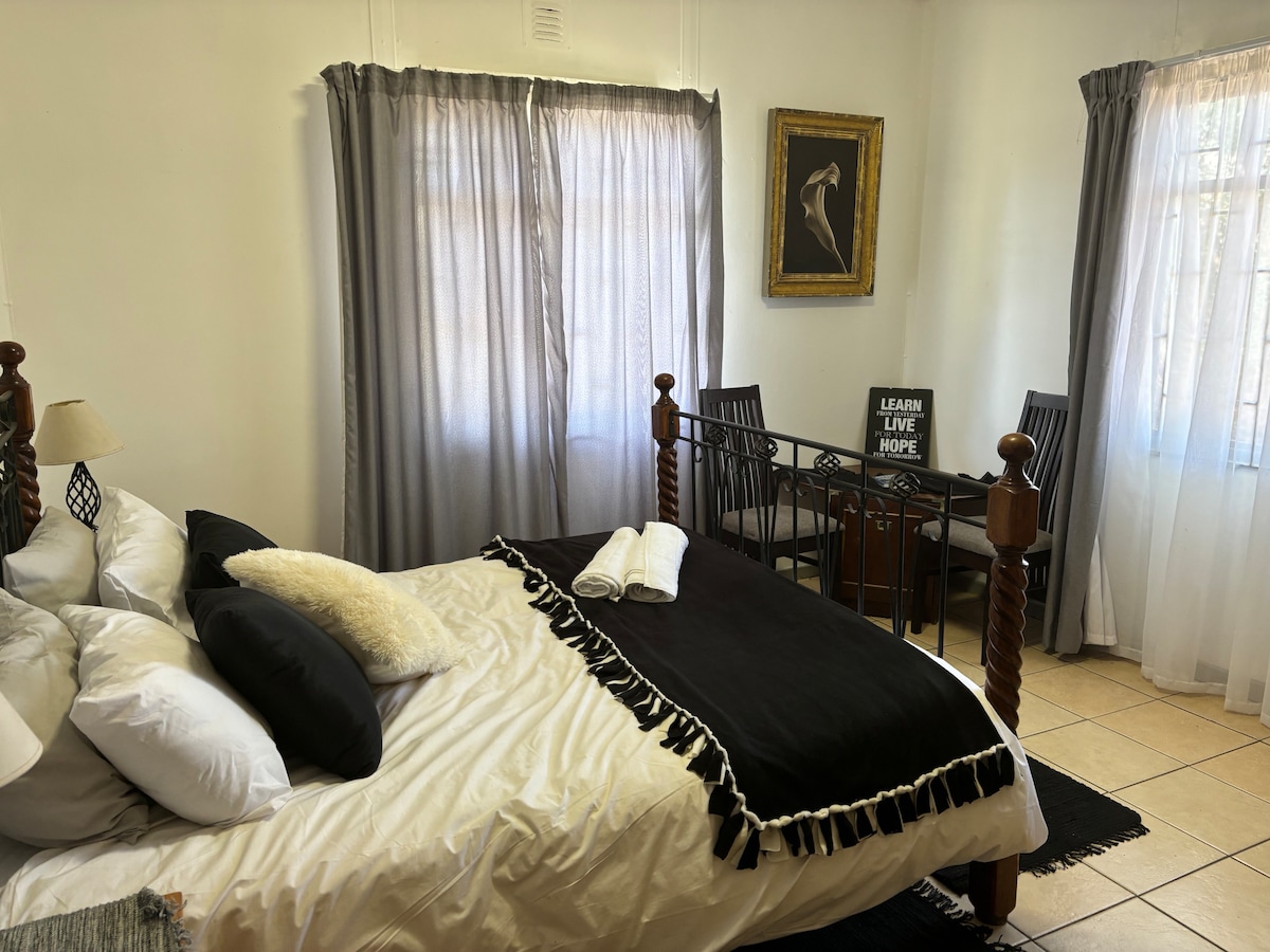 A sleeping area features a spacious bed with an assortment of pillows. A black and white throw blanket is draped over the bed. Natural light floods in through sheer curtains, illuminating a small table and chairs positioned near the window.