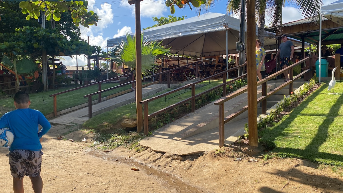 A pathway leads to a shaded outdoor dining area, framed by greenery and structures. Ramps provide accessibility, while a child stands on the path, holding a soccer ball. Tables and chairs are arranged under large canopies, suggesting a lively atmosphere.