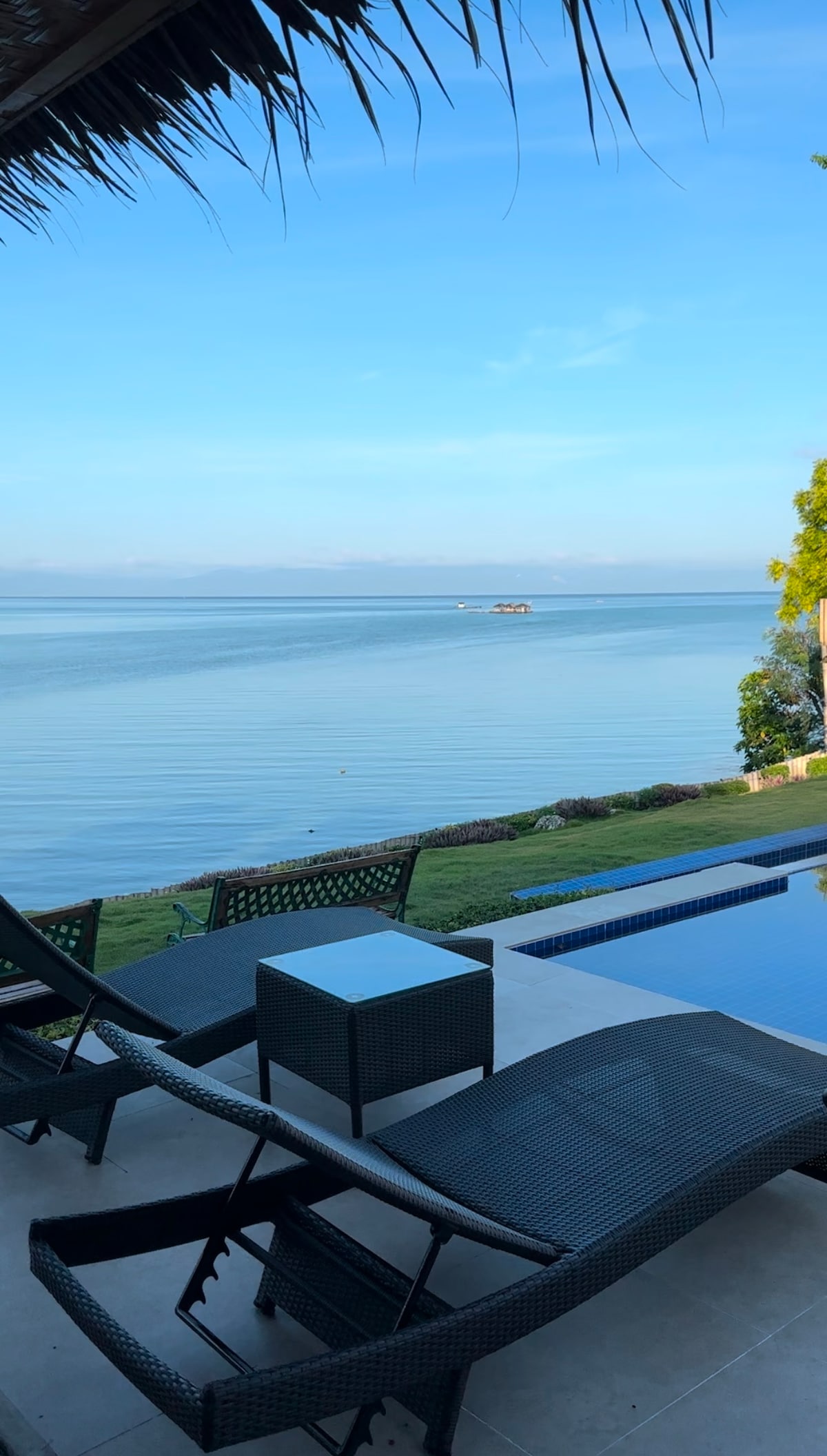 A sunny outdoor area features two loungers positioned beside an infinity pool, overlooking a calm sea. Lush greenery and a skyscape complement the tranquil scene, which includes a distant boat and the gentle reflection of clouds on the water's surface.