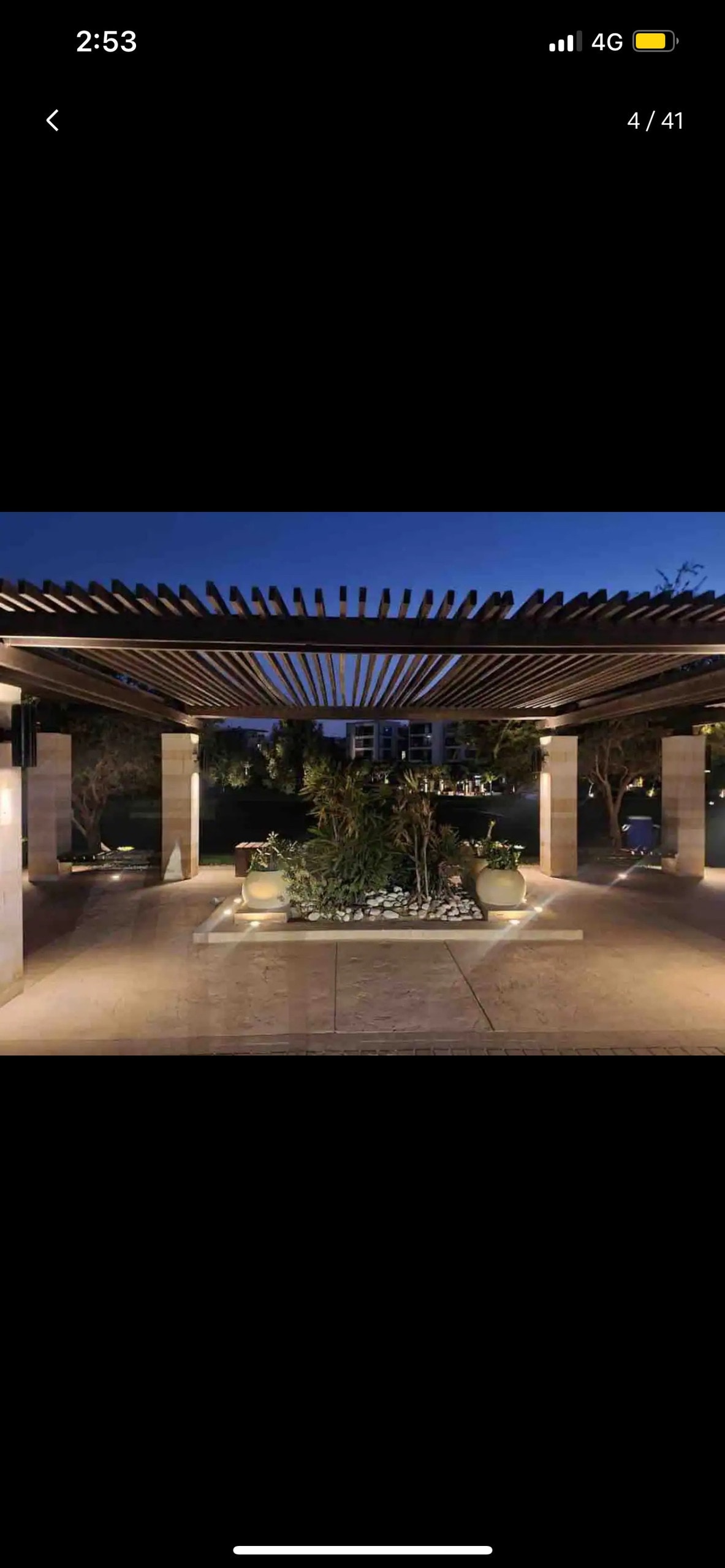 An outdoor area is highlighted by a wooden pergola, casting gentle shadows over the stone pathway. Planters with lush greenery and soft lighting create a serene ambiance as night falls, inviting relaxation in this tranquil setting.